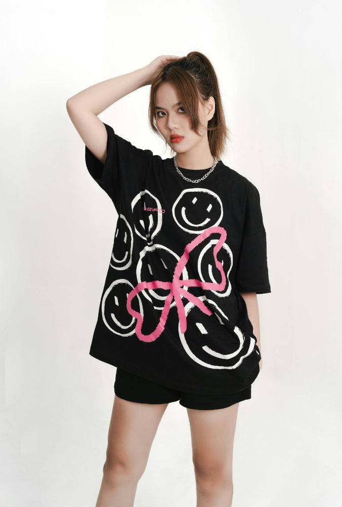 Femme portant un t-shirt oversize noir avec motifs smiley blancs et un grand dessin rose stylisé.